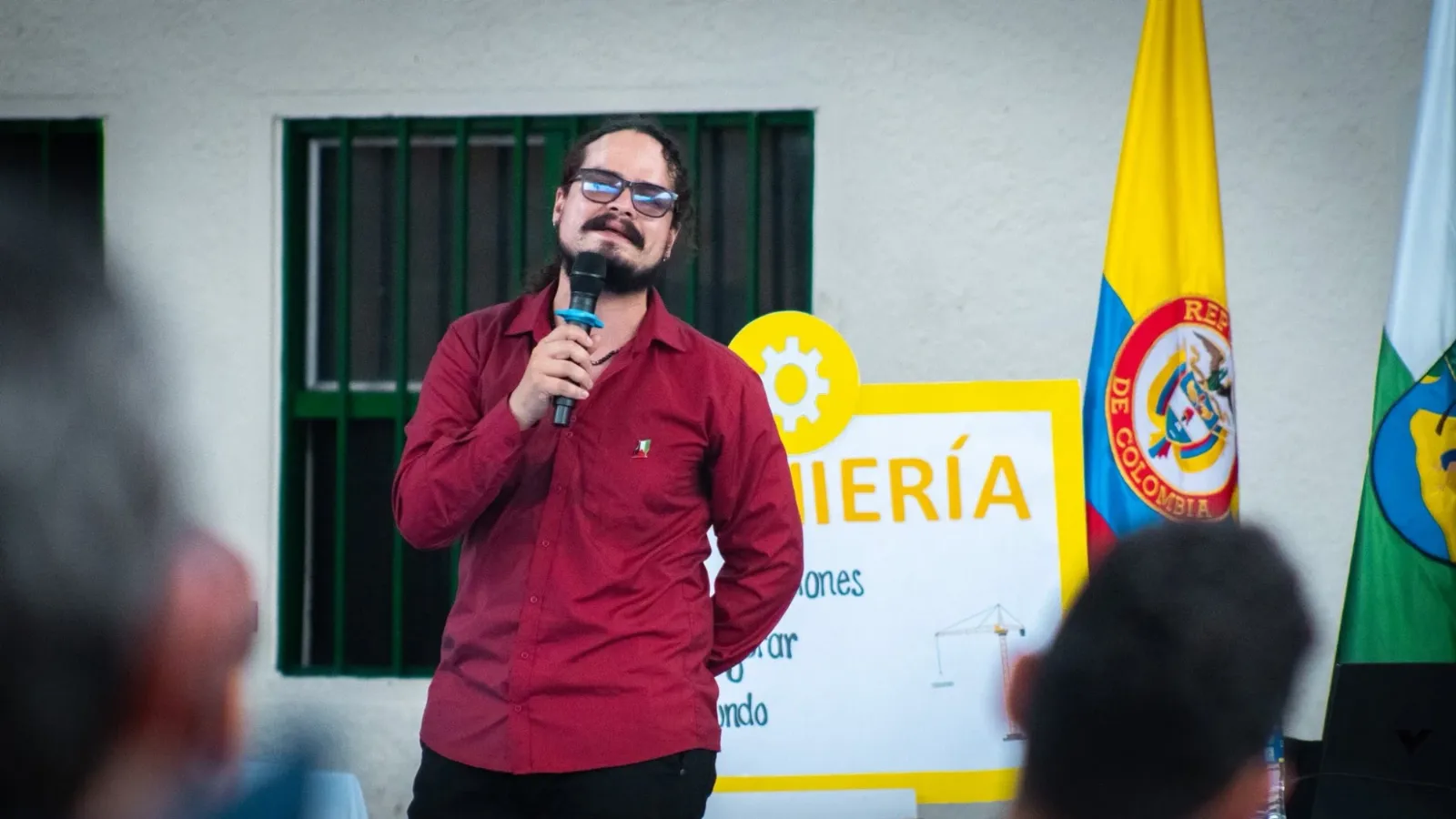 Persona hablando al micrófono frente a un letrero amarillo y una bandera de Colombia.
