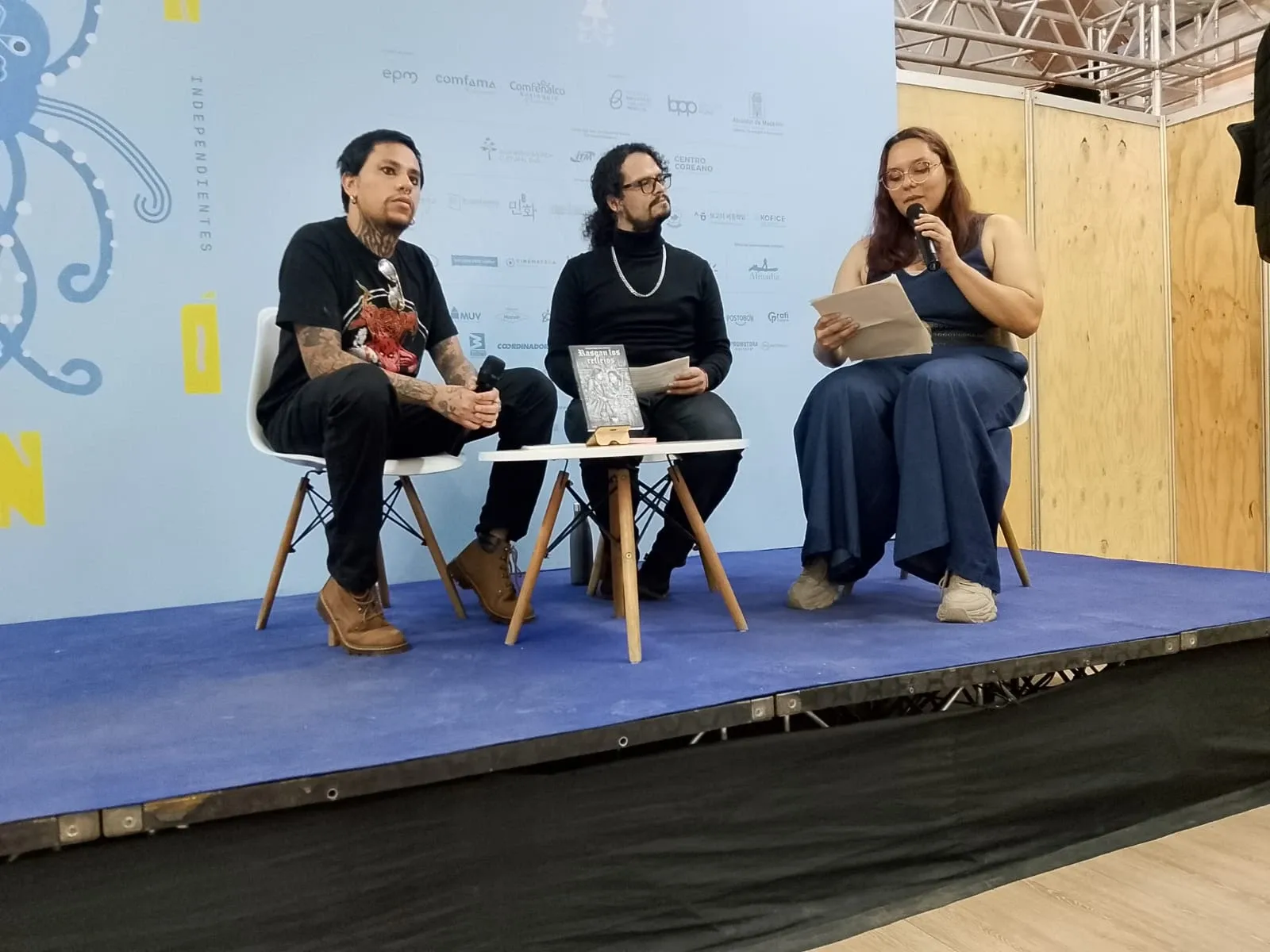 Tres personas sentadas en el escenario de la Fiesta del Libro de Medellín, junto a una mesa con el libro Rasgan los reflejos.
